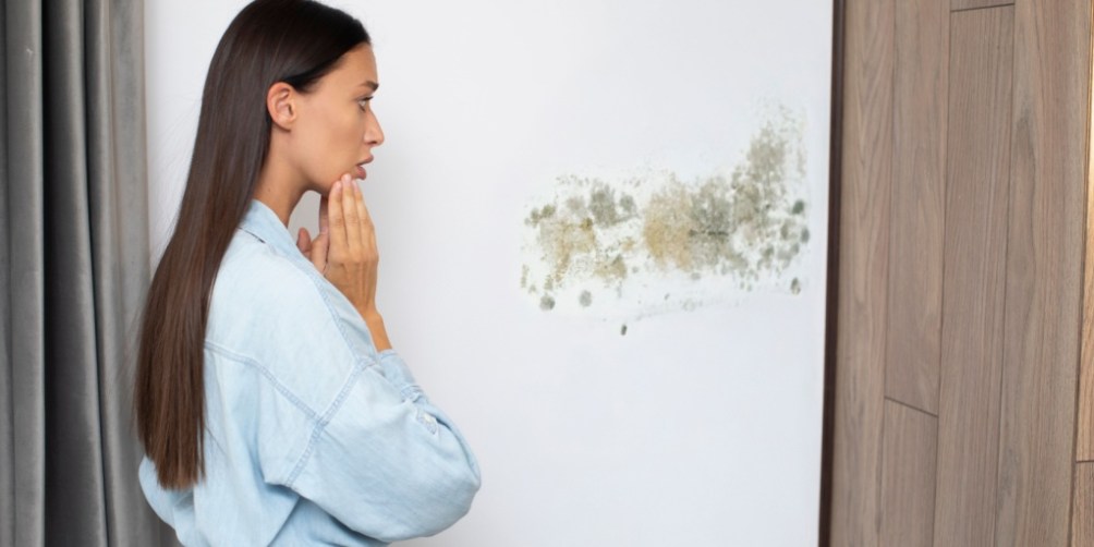 A concerned woman stands in front of a white wall covered in mold spots, reflecting on whether renters insurance covers mold damage in rental properties.