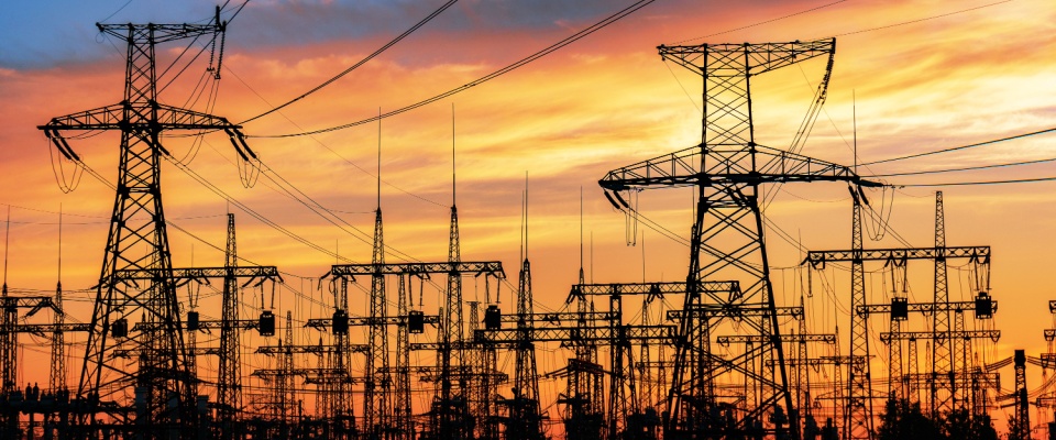 A network of high-voltage electricity transmission towers and power lines silhouetted against a vibrant sunset sky, visually representing the infrastructure used for various electricity plans.