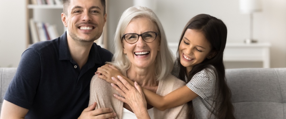 A senior woman sits on a comfortable sofa embraced by her family in a bright living room, looking to downsize to be closer to her family.