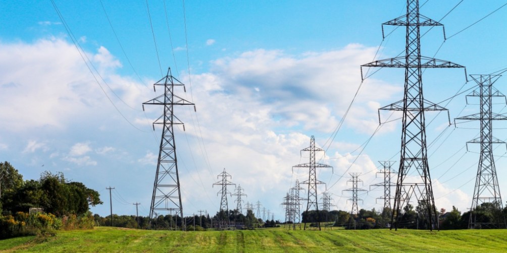 A row of tall electricity transmission towers stretches across a green field under a bright blue sky, illustrating a step in the process of how electricity works.