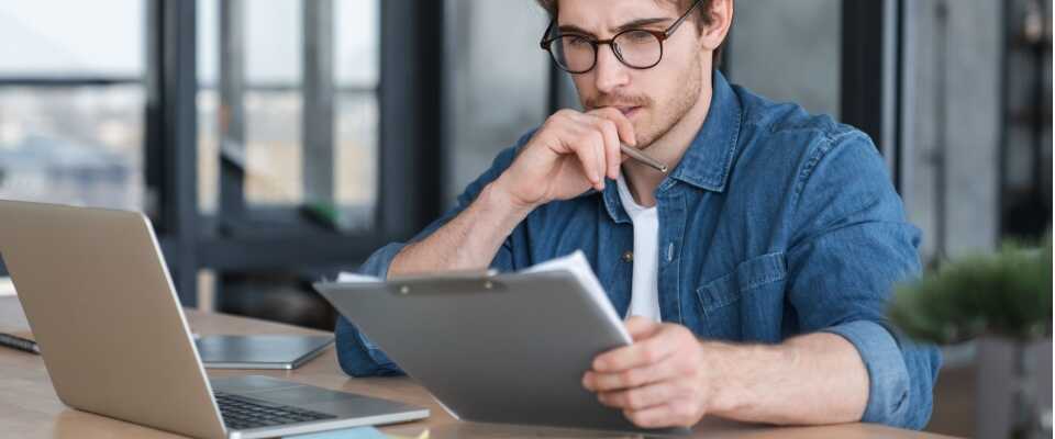 Man wearing glasses sits at a desk with a laptop, intently reading the renters insurance policy on a clipboard, looking into changing the policy's effective date.