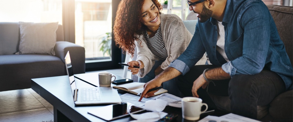 Smiling couple sitting in their apartment reviewing bills and paperwork on a laptop, preparing proof of renters insurance for move-in.