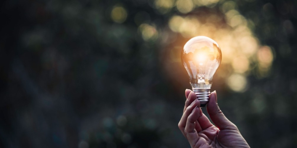 Hand holding a glowing light bulb against a natural background, representing the transformative power of the invention of electricity.