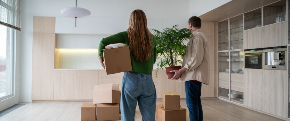 Couple carrying moving boxes and a houseplant into a bright, modern apartment after securing one of the available month-to-month rentals in their area.